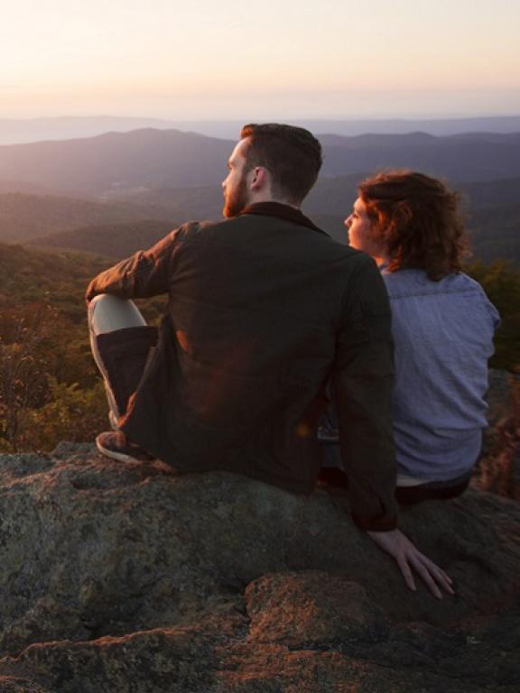 Two serene-looking people sit side-by-side on large rocks overlooking the hills and forests of Shenandoah National Park in Virginia, USA at sunset. The glow of the sun reflects on their faces.